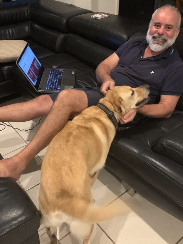 A smiling man with a grey beard is sitting on a leather couch patting a Labrador